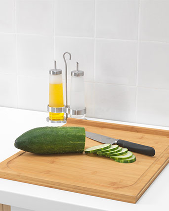 Un pepino verde medio cortado y un cuchillo de mango negro reposan sobre una tabla de cortar de bambú marrón claro. Dispensadores de aceite y vinagre de acero inoxidable cerca.