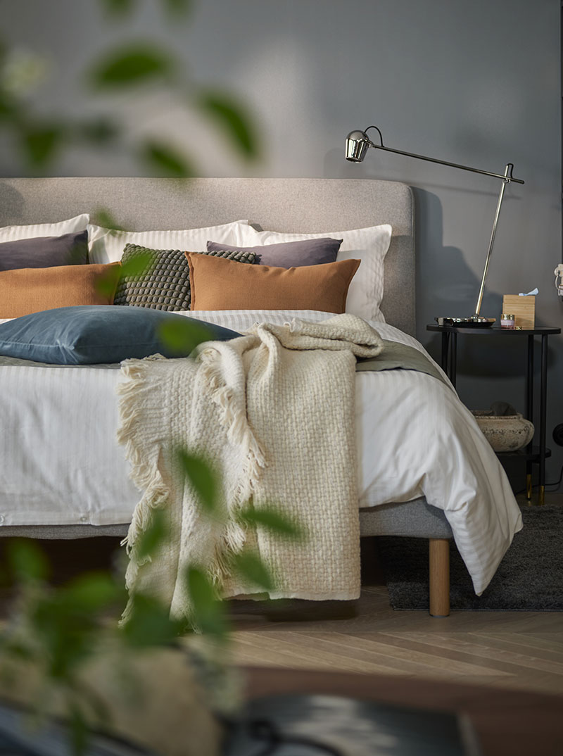 Vista frontal de una cama con edredones mullidos y muchas almohadas y cojines, hay una mesilla de noche con una lámpara de mesa de metal con bisagras.
