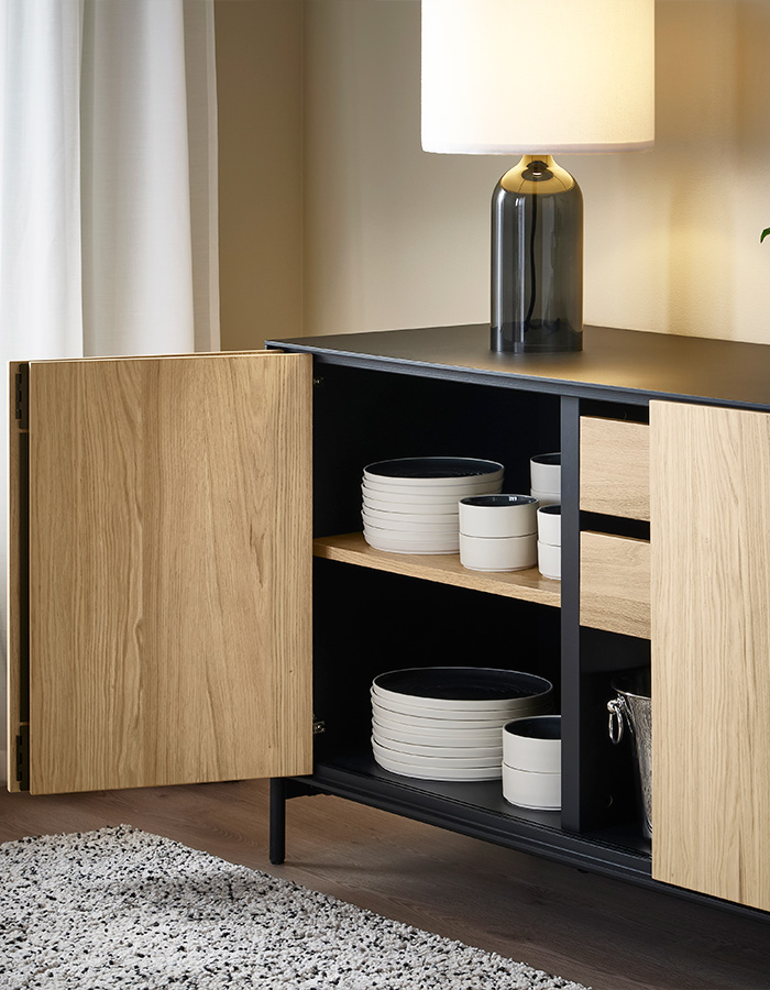 A modern black and wood sideboard with open doors reveals white and black dishes. A stylish table lamp with a dark base sits on top.