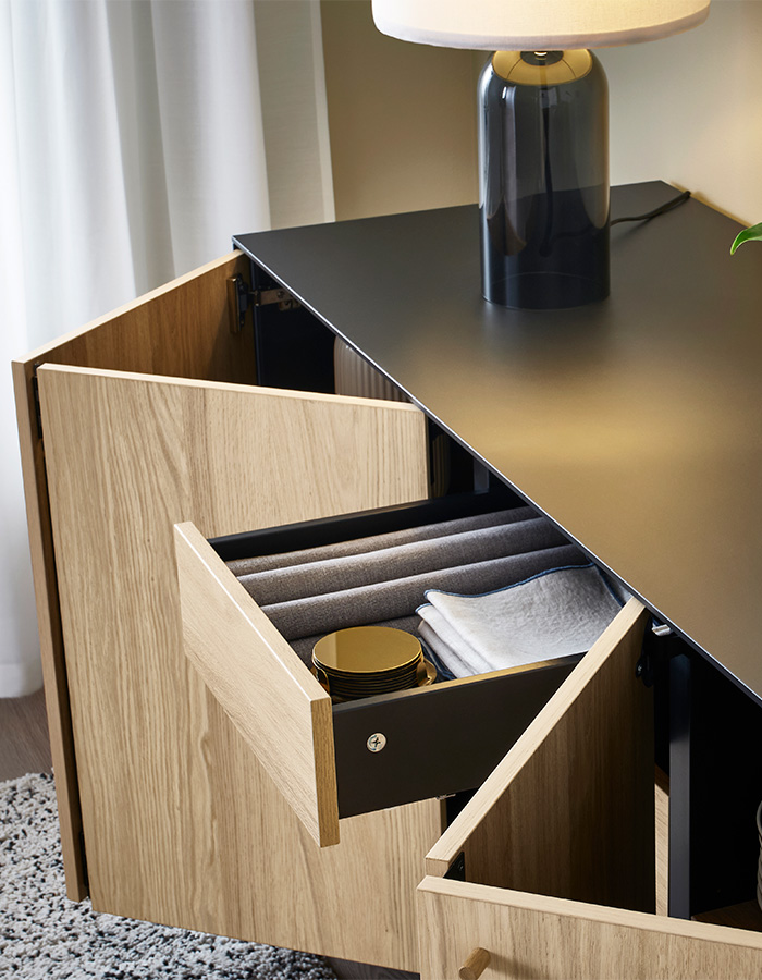 A modern black and wood sideboard with a unique open drawer revealing a gold pot and white cloth. A dark-based lamp sits on top.