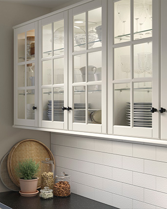 Upper kitchen cabinets with white glass doors and black knobs, displaying dinnerware and glassware, above a countertop with jars and decorative plants.