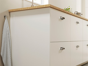 Close-up of matte white lower kitchen cabinets with metal handles and light wood countertop, with a dish towel hanging on the side.