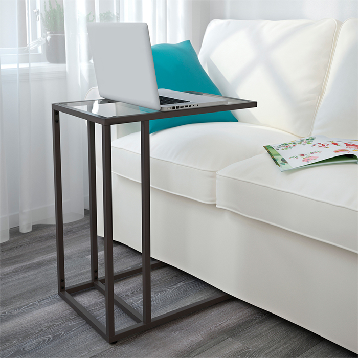 A dark metal C-table with a glass top holds a silver laptop beside a white sofa with a bright turquoise pillow and open book.