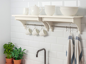 Two wall shelves in a kitchen holding light-colored kitchen towels, cups, bowls, and a teapot; below is a sink with a faucet.