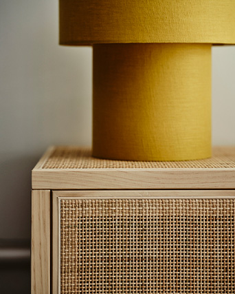 A modern yellow lamp sits on a light wood cabinet with a woven rattan front.