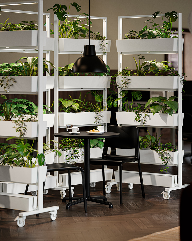 A café-style break room with MITTZON frames filled with plants, surrounded by black STENSELE tables and chairs in the centre.