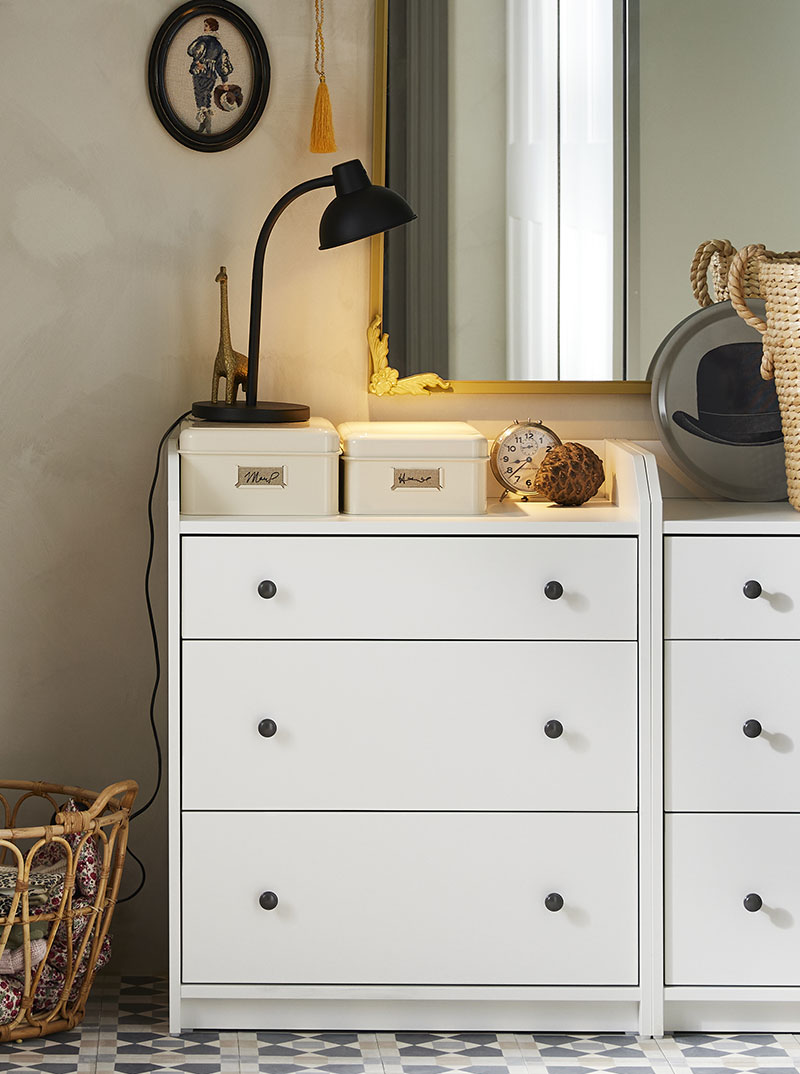 Three-drawer chest with metal storage boxes, lamp, and a partially visible basket on the floor in a rustic setting.