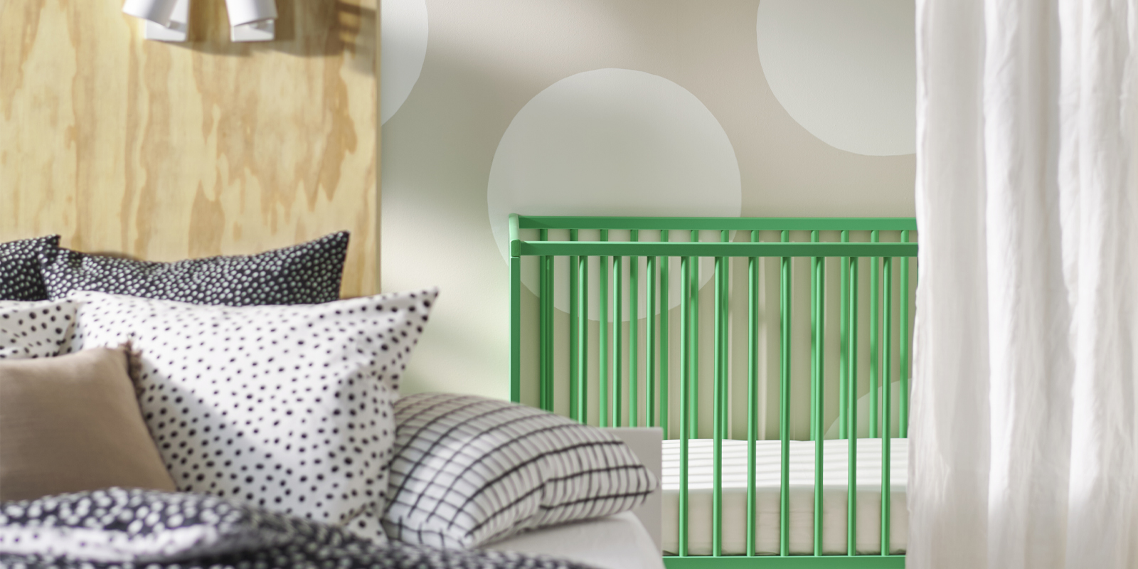 A green GULLIVER cot is placed against a lightcoloured polka-dot wall. A bed covered in BJÖRKGRÅMAL bed linen is in front.