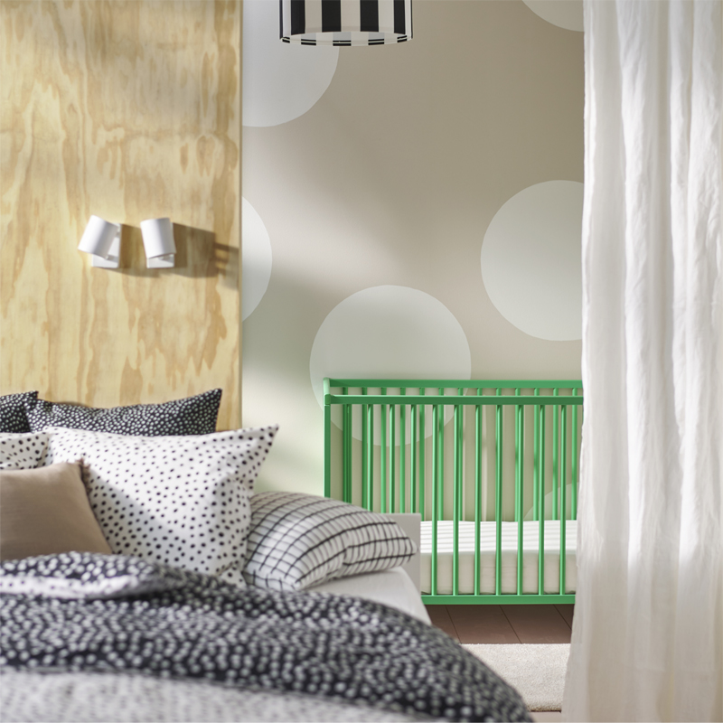 A green GULLIVER cot is placed against a lightcoloured polka-dot wall. A bed covered in BJÖRKGRÅMAL bed linen is in front.
