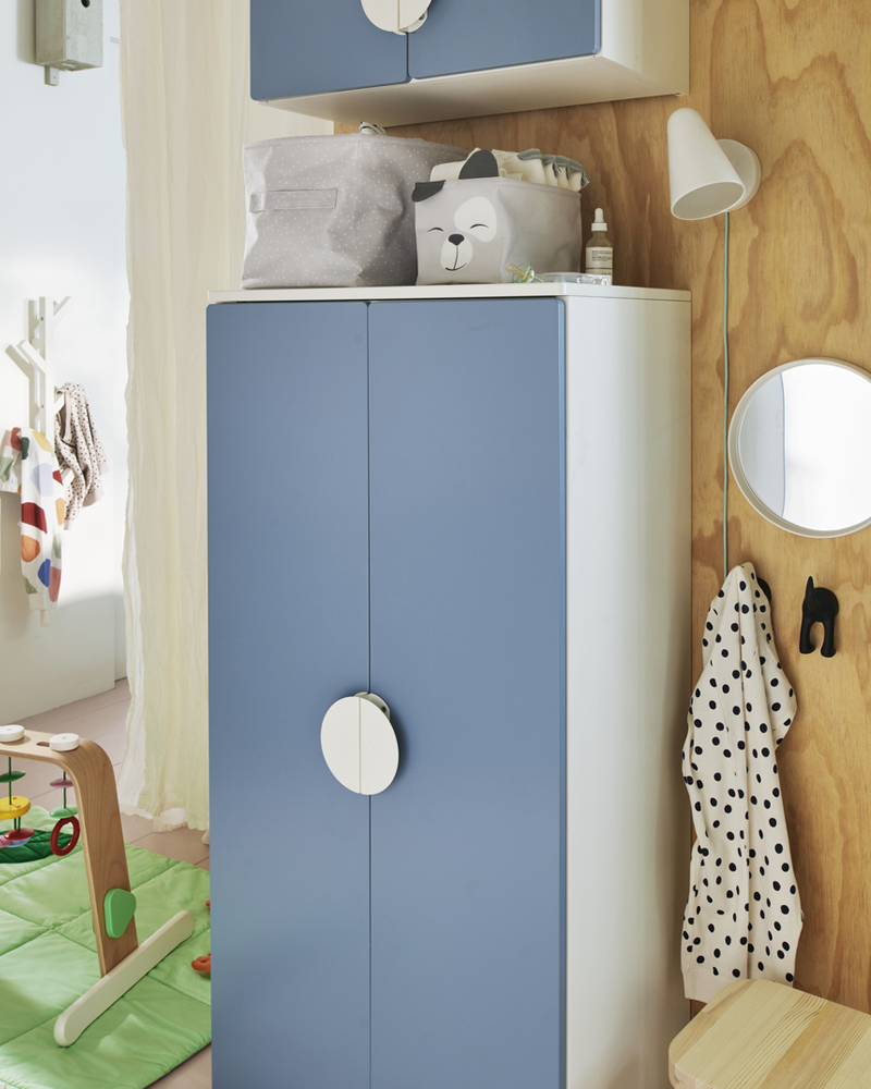 A blue SMÅSTAD wardrobe against a wooden wall with two storage boxes holding baby things on top and a wall light beside it.