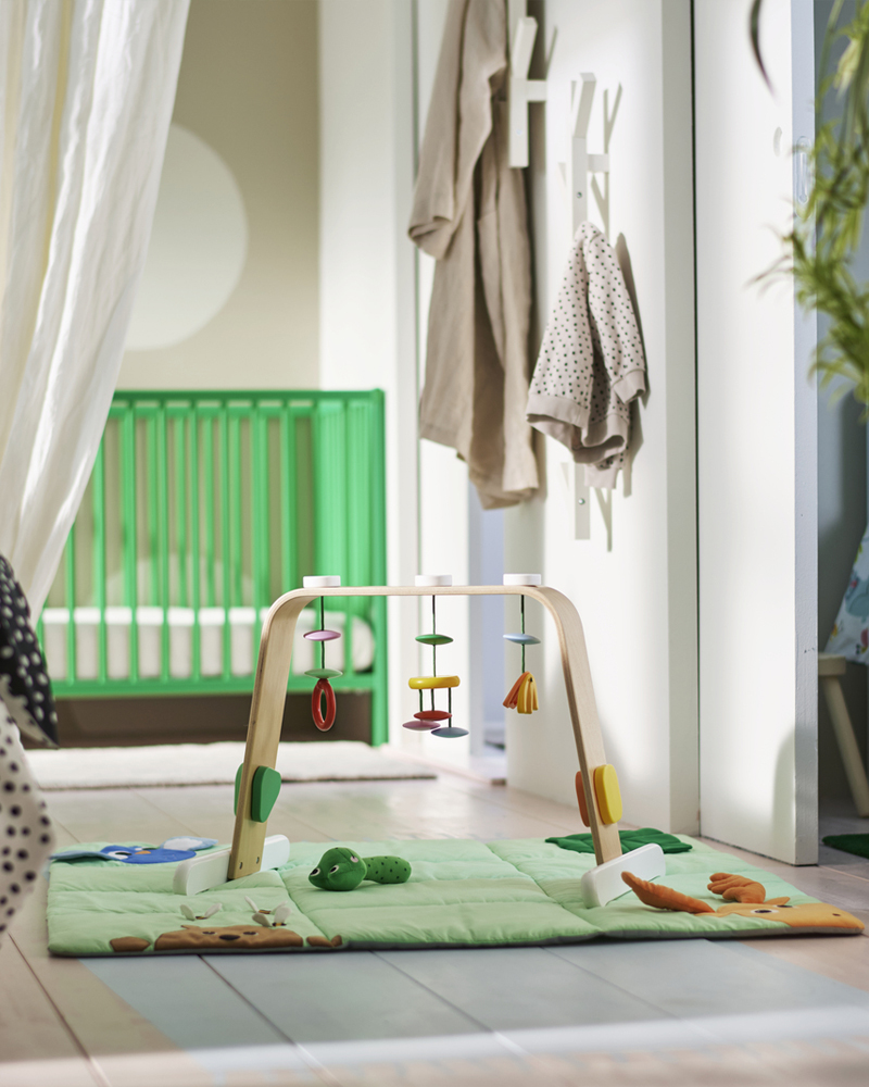 A multicoloured LEKA baby gym with hanging toys on a green playmat on the floor in a bedroom with a green GULLIVER cot.