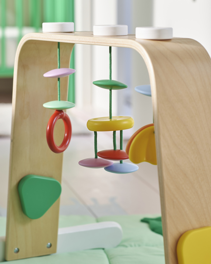 A multicolour LEKA baby gym with bright coloured hanging rings and shapes on top of a green playmat in a baby’s bedroom.