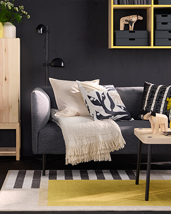 Salón IKEA acogedor con sofá gris, cojines y manta. Soluciones de almacenaje con estantería amarilla para un espacio funcional y con estilo.