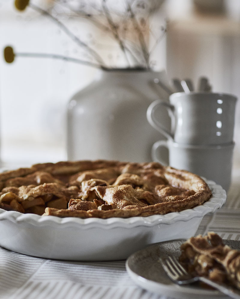 Sobre un mantel a rayas, rodeado de cubiertos y jarrones, una tartera blanca de HÖSTAGILLE rellena de tarta de manzana.