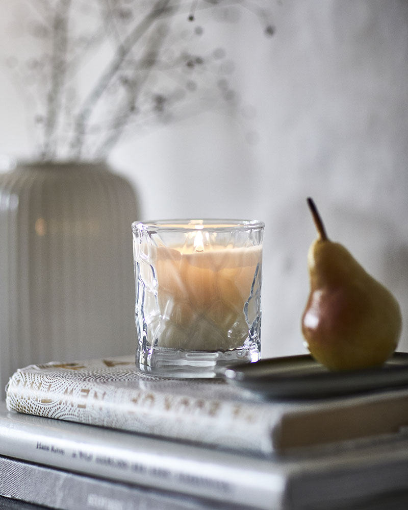 Una vela perfumada HÖSTAGILLE en un tarro de cristal se coloca sobre libros apilados junto a un jarrón decorativo y una pera