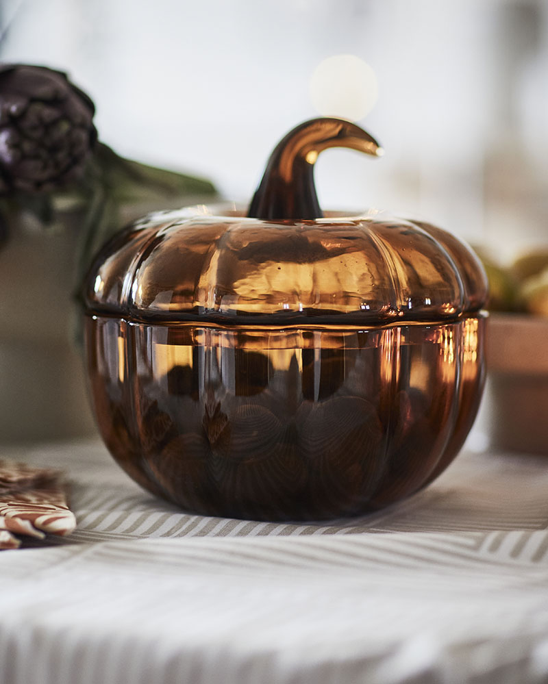 Primer plano del cuenco decorativo HÖSTAGILLE con tapa, de color marrón amarillento, con un diseño en forma de calabaza sobre una mesa estampada