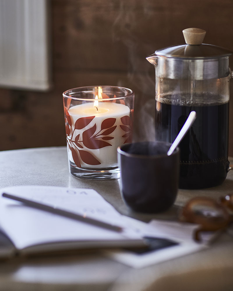 En una mesa, junto a una prensa francesa y una taza humeante, hay una vela aromática HÖSTAGILLE en un vaso.