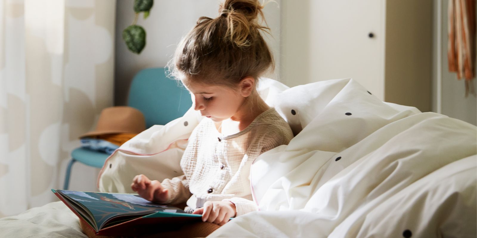A girl reading on a bed with a quilt around her, with a dotted white/pink VÄNKRETS quilt cover, and two white pendants above.