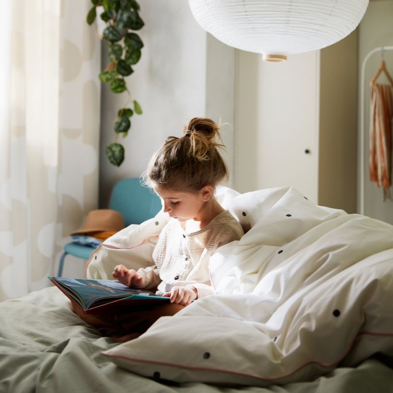 A girl reading on a bed with a quilt around her, with a dotted white/pink VÄNKRETS quilt cover, and two white pendants above.
