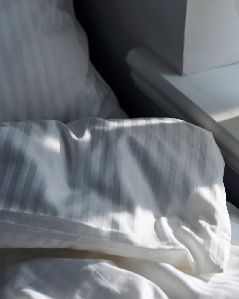 The corner of a folded-over duvet covered in NATTJASMIN bed linen in white in a sunny space by a shelf.