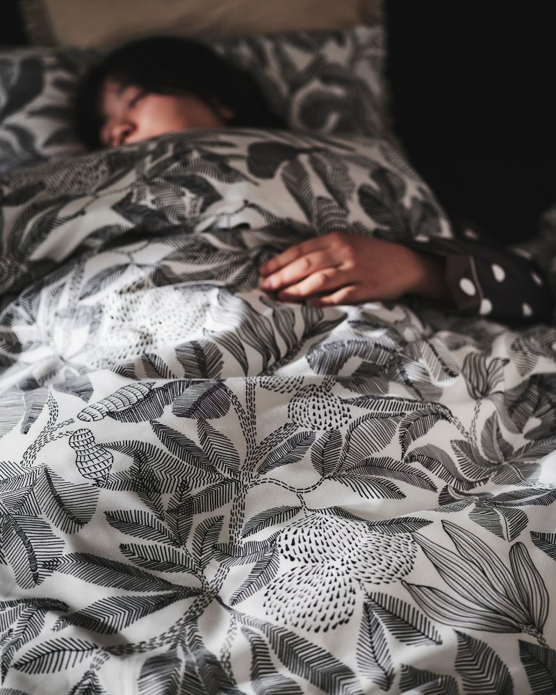 A person asleep in a SLATTUM upholstered bed under a duvet that’s covered in black and white BROKDRACENA bed linen.