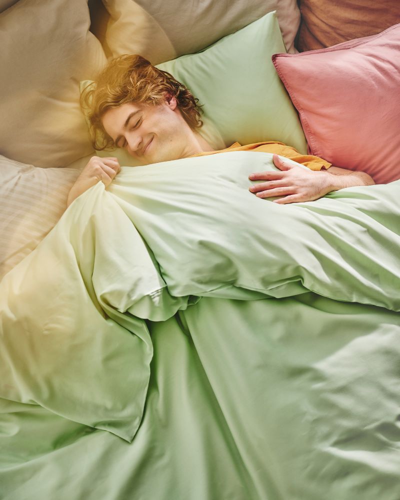 A person smiles as they lie in bed covered with light green PILTANDVINGE bed linen, with other pillows around them.