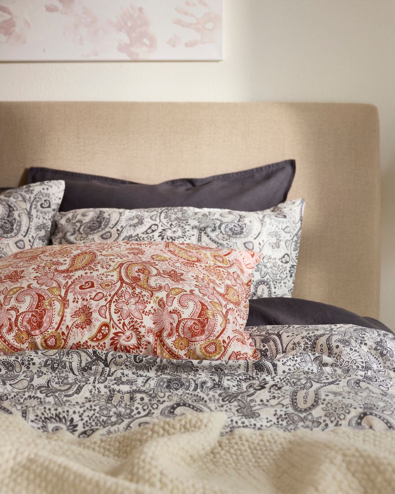 The top of a bed that’s covered with two types of RODGERSIA paisley pattern bed linen, one in grey and the other in red.