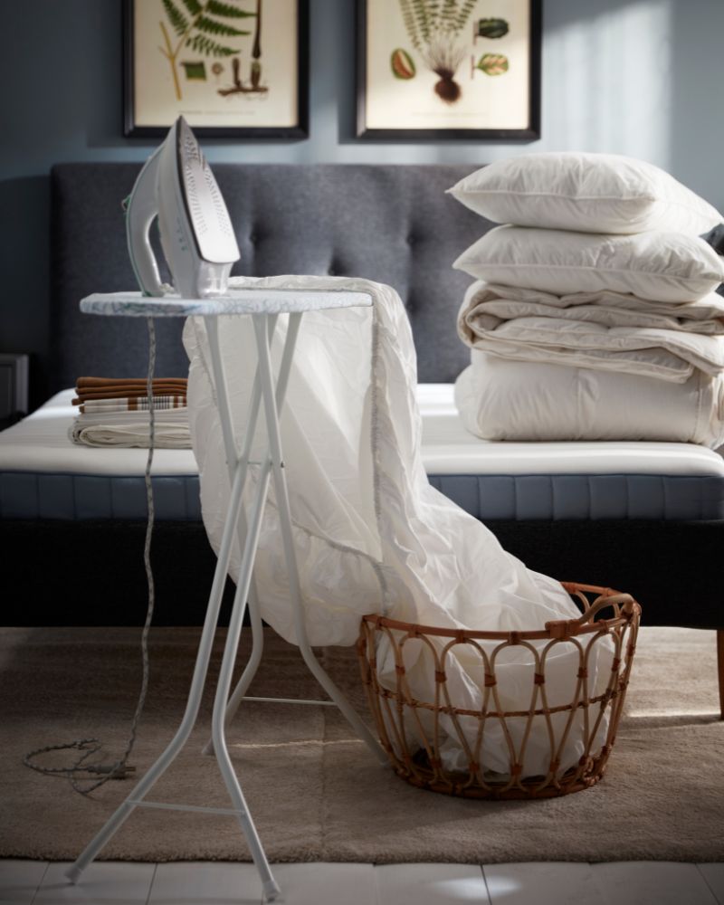 A white NATTJASMIN fitted sheet that is half in a baske and half laid out on a RUTER ironing board in front of an unmade bed.