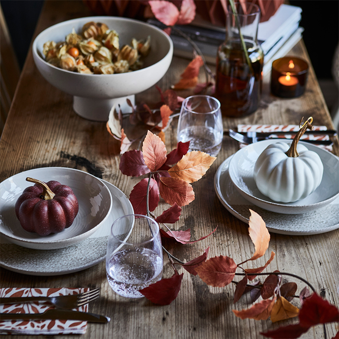 Rustic wooden table set for an autumn dinner with ceramic plates, white and burgundy decorative pumpkins, water glasses, cutlery, and reddish-brown autumn leaves, with a lit candle in the background.