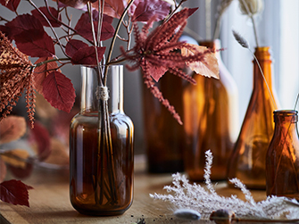 Close-up of an amber glass vase with reddish decorative leaf branches, accompanied by other blurred amber glass bottles and vases in the background, creating a warm and autumnal atmosphere.