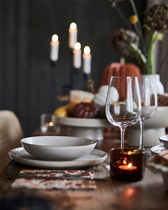 Detail of an autumn table setting with a textured ceramic plate, an empty wine glass, a patterned placemat, and a lit candle in a dark glass holder, with decorative pumpkins and a candelabra in the background.