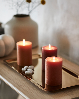 Three lit cylindrical candles in orange and burgundy tones, placed on a gleaming golden tray with small decorative white pebbles, creating a cozy and warm ambiance.