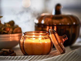 Lit wax candle inside a smoky glass container with a matching lid, near cinnamon sticks and a bowl of physalis, all with a soft blurred background suggesting an intimate atmosphere.