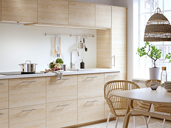 Modern light wood kitchen with white countertop, utensils, and natural lighting.