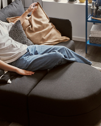Detail of a sofa with light-toned pillows and cushions. A white wall lamp above the headboard illuminates the area, with a bedside table to one side.