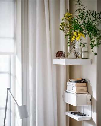 Room corner with white floating shelves and light curtains. The shelves hold vases with plants, books, and a phone, organizing vertical space.