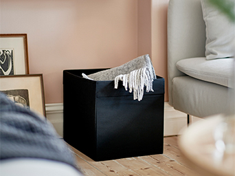 Cesto de almacenaje negro de IKEA en un salón, con una manta gris. Solución versátil para organizar mantas o cojines, perfecta para el hogar.