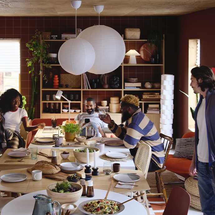 Mesa de comedor de madera clara en un salón con estantería. Lámparas de papel colgantes. Ambiente social con gente comiendo.