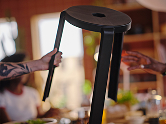 Un taburete de madera oscura con asiento hexagonal es sostenido en el aire. Ambiente de comedor desenfocado de fondo.