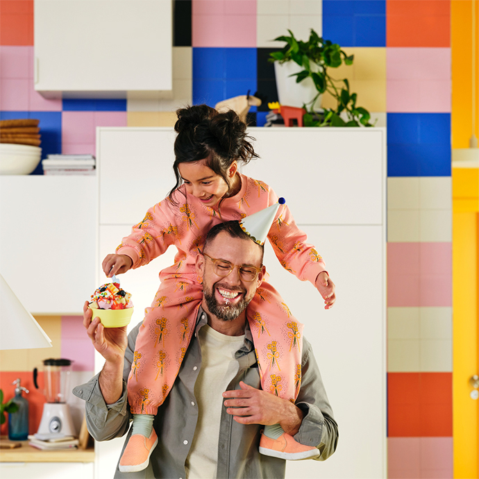 Un padre sonriendo lleva a su hija en los hombros en una cocina de colores. La niña sostiene un bol de fruta.