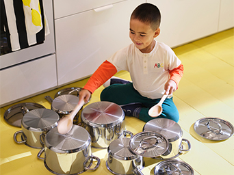 Un niño sentado en el suelo amarillo de una cocina, rodeado de ollas y sartenes metálicas. Sostiene dos cucharas de madera.