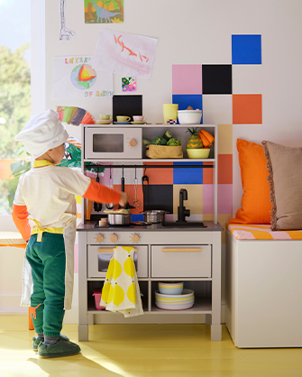 Un niño vestido de cocinero juega con una cocinita de madera de color gris claro. Hay dibujos en la pared y un banco con cojines.