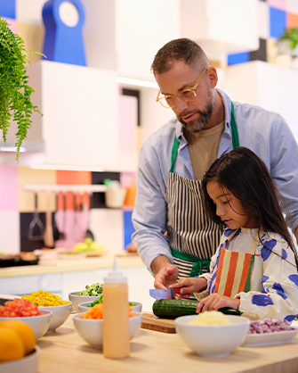 Un padre y su hija cortan pepino en una cocina con azulejos multicolores. Varios boles de ingredientes sobre la encimera de madera.