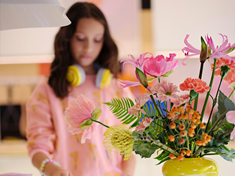 Un ramo de flores rosas, anaranjadas y amarillas en un jarrón. Una niña con auriculares y una sudadera rosa aparece borrosa de fondo.