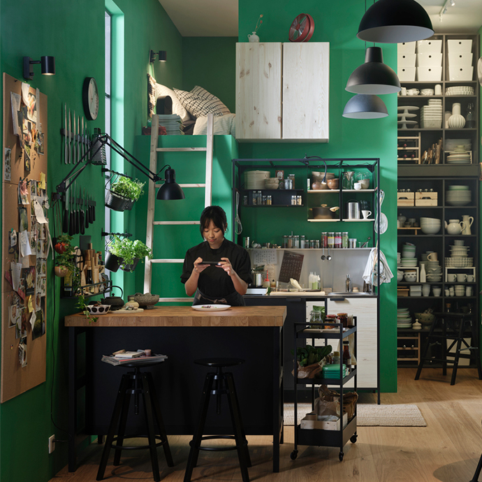 Una mujer en una cocina de color verde intenso con una cama en un altillo. Hay una mesa de isla con taburetes.