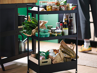 Carro auxiliar de tres niveles en color negro, con alimentos y productos. Cerca, una tetera y un mueble con cajones blancos.