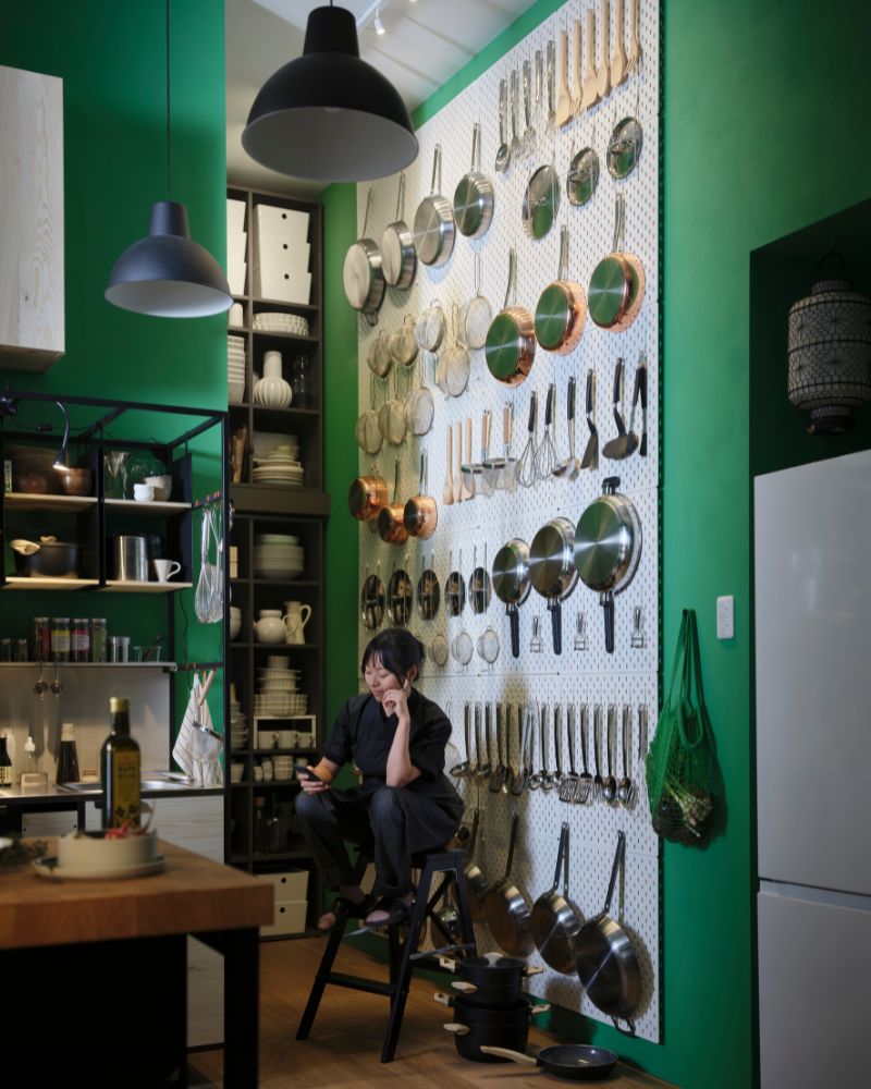 A person is sitting on a stepladder in front of a wall adorned with SKÅDIS pegboards, with kitchen items attached to them. 