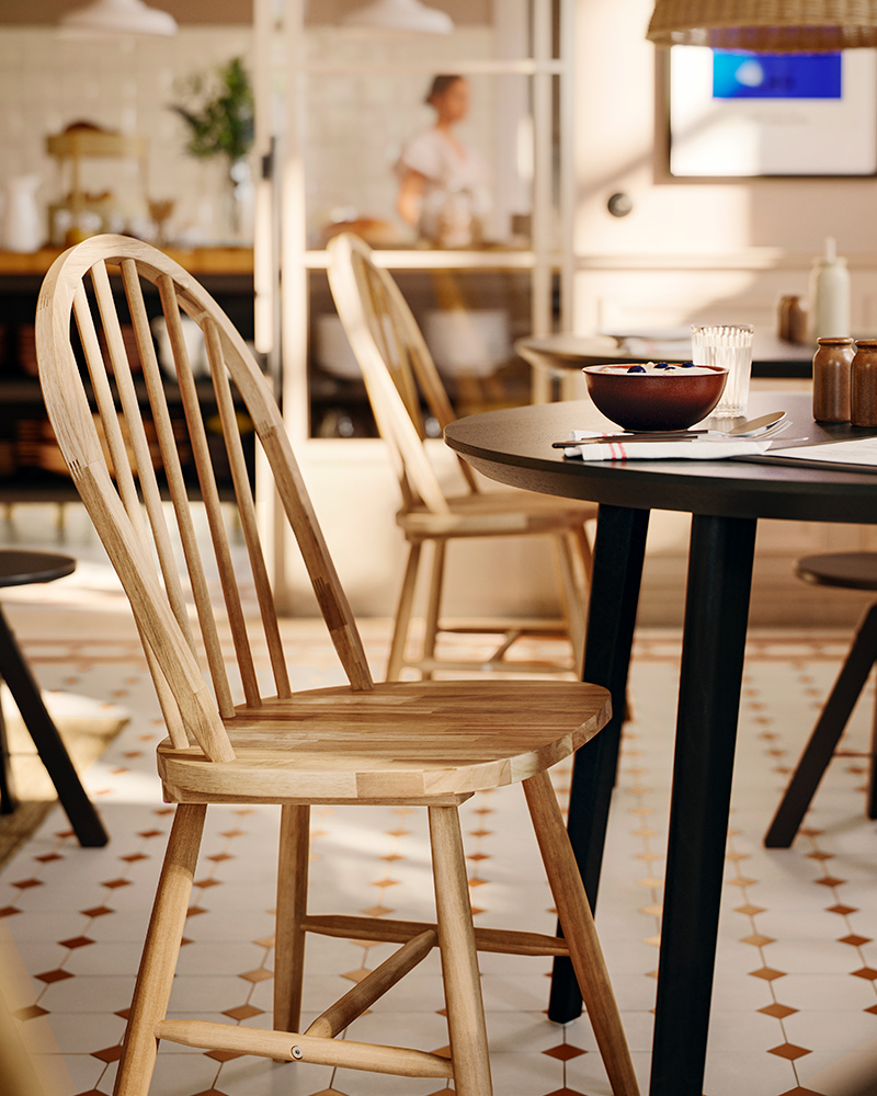 A solid acacia SKOGSTA chair with an arched backrest and spindles is at a dark, round table in a commercial dining area.