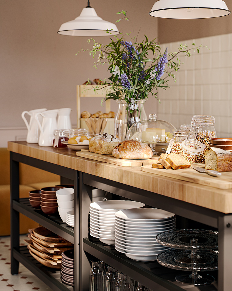 Two VADHOLMA kitchen islands with oak veneer worktops and black shelves hold a breakfast buffet on top and dinnerware below.