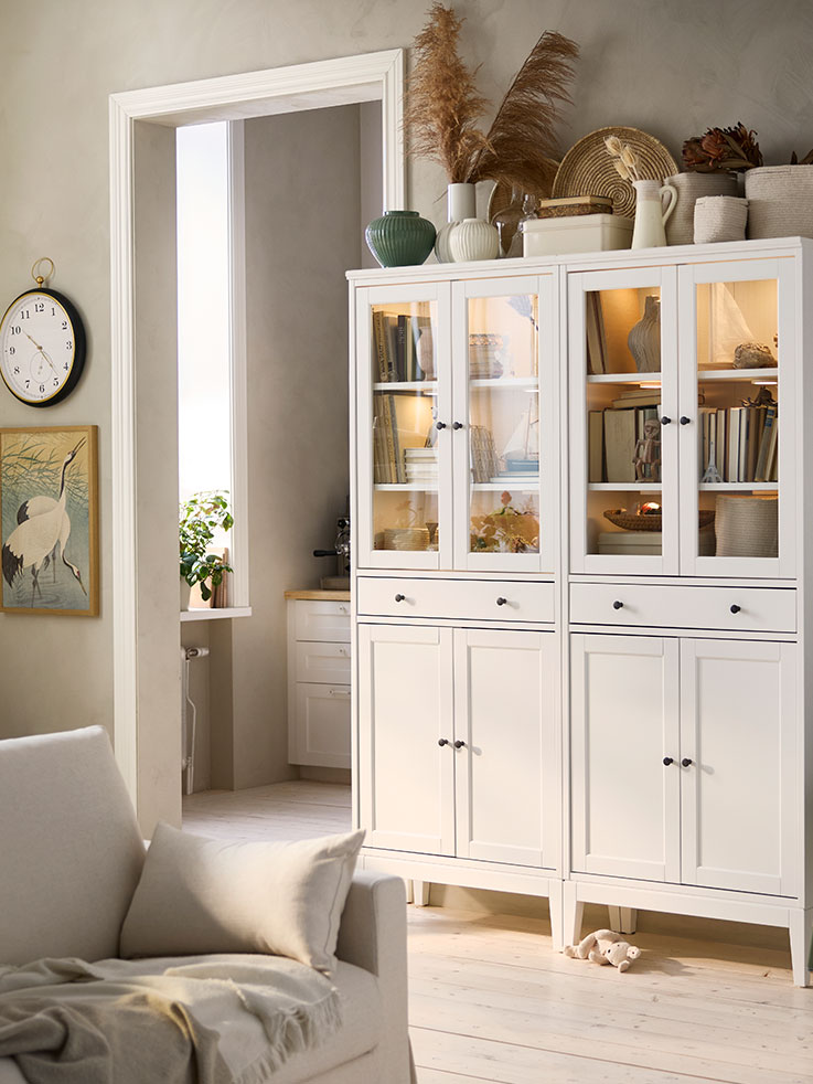 Dos armarios altos IDANÄS blancos con puertas de cristal y 1 cajón, colocados uno al lado del otro, tienen libros y recuerdos guardados en su interior.